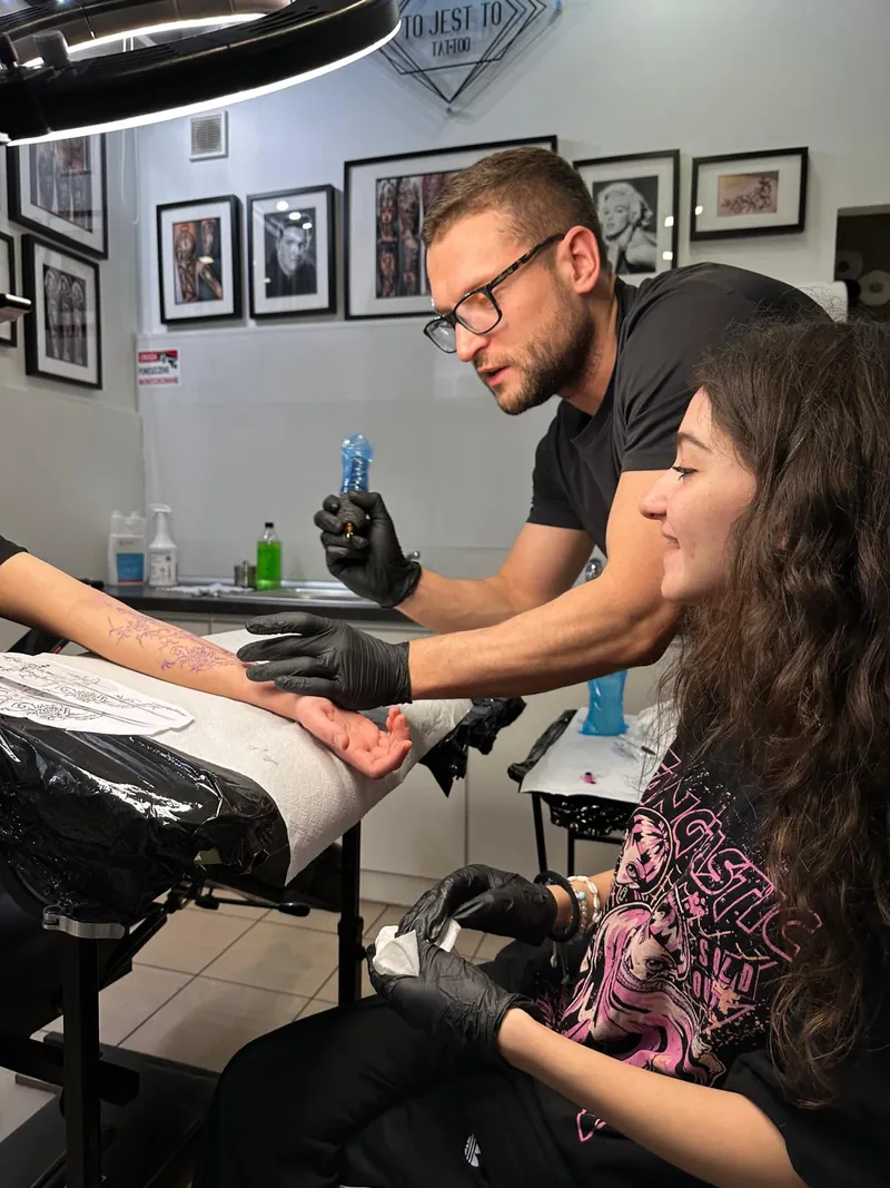 Kurs tatuażu w&nbsp;To Jest To Tattoo: mężczyzna w&nbsp;rękawiczkach pokazuje siedzącej obok osobie przygotowany wzór na&nbsp;przedramieniu, obok lampa pierścieniowa i&nbsp;ściana ze&nbsp;zdjęciami prac.