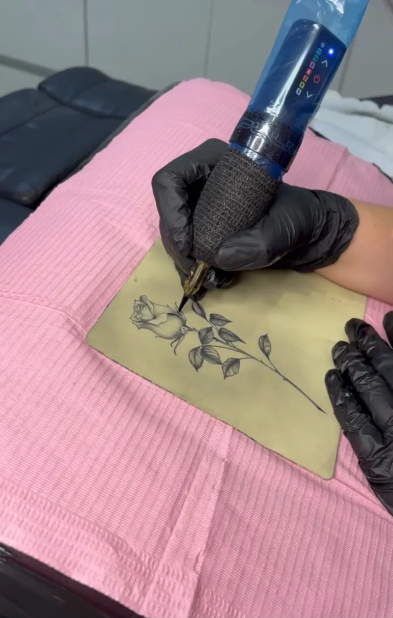 Kurs tatuażu w&nbsp;To Jest To Tattoo: dłoń w&nbsp;czarnej rękawiczce prowadzi maszynkę do&nbsp;tatuażu po&nbsp;sztucznej skórze z&nbsp;wzorem róży, na&nbsp;różowym podkładzie.