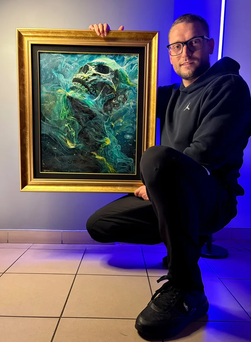 Artysta kucający z&nbsp;obrazem w&nbsp;złotej ramie, obraz przedstawia czaszkę w&nbsp;turkusowo-złotych barwach, obraz z&nbsp;pracowni To Jest To Tattoo
